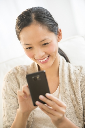 Happy Asian mid adult woman using mobile phone while relaxing on sofaの写真素材