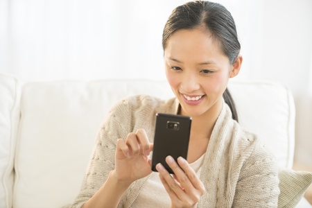 Happy Asian mid adult woman using smart phone while relaxing on sofaの写真素材