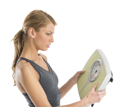 Worried young woman in sports clothing looking at weight scale against white backgroundの写真素材