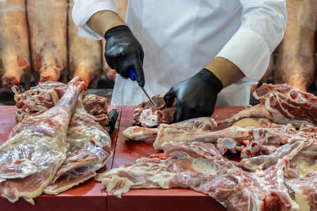 Butcher man in black gloves is cutting lamb meat in butcher shop. Chef cook with white apron is cutting meatの写真素材
