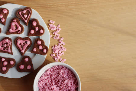 Heart shaped chocolate cookies with pink sugar glaze and sprinkles on a plate on wooden table. Valentine's day backgroundの写真素材
