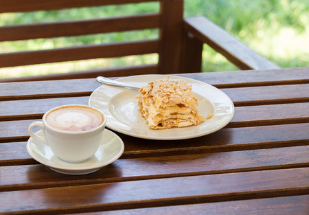 Napoleon cake, cappuccino coffee on the wooden table on the veranda in the cafe, in the gardenの写真素材