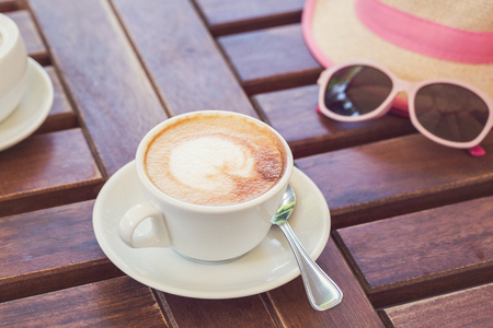 On a wooden brown table is a cup with cappuccino, a hat, glasses. Summer, cafe, rest. Toning vintageの写真素材