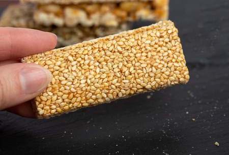 Female hand holding a granola bar in her hand. Healthy sweet dessert snack. Sesame, hazelnut, sunflower in honey. Gozinaki is Georgian national food, oriental sweet on black stone backgroundの写真素材