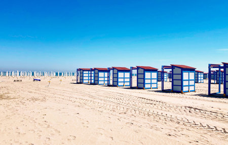 Chioggia, Italy - May 11, 2025: Vibrant beach huts are arranged on sandy beach with clear blue sky,...