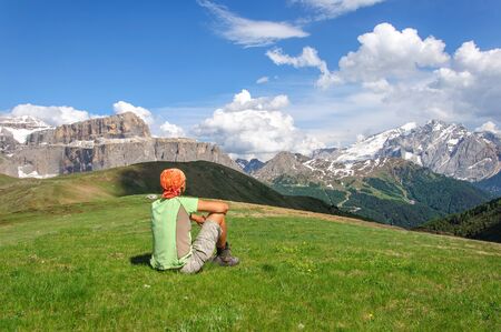 Young man sitting on a green lawn and are looking at snow-covered dolomites, Italy.の写真素材