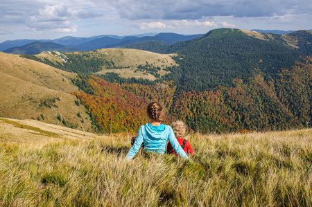 Happy mom and daughter sit on a mountainside in the Carpathians and admire the autumn forestの写真素材