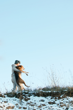 young man carries a dog on the slope of a winter hill covered with snowの写真素材