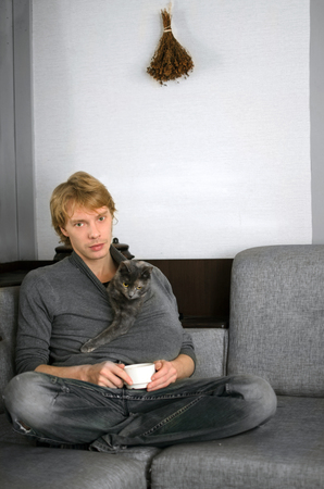 young man in a gray sweater with a Cup of coffee in his hands and his gray catの写真素材