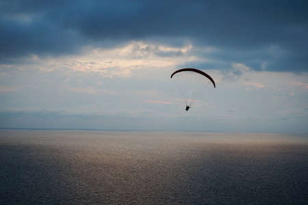 a paraglider flies over the sea at sunset against the background of the light part of the sky between the dark sea and dark cloudsの写真素材