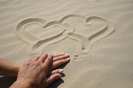 a man's hand gently touches a beautiful hand with a well-groomed manicure of his beloved. Their hands lie on the sand of the sea beach on which two hearts are drawn with a fingerの写真素材