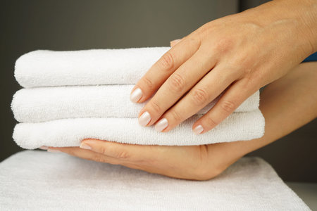 woman with beautiful hands with good manicure holds a neatly folded stack of white terry towels indoorsの写真素材