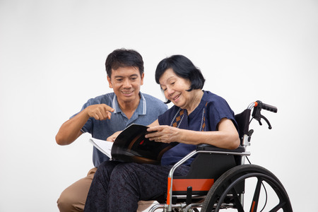 Elderly woman reading a book on wheelchair with her son take care.の写真素材
