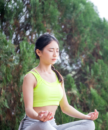 Beautiful young woman wearing neon green aerobic clothing and grey aerobics pants enjoying yoga outdoors. Resting after doing yoga exercises, sitting in ardha Padmasana, Lotus pose, relaxing.の写真素材