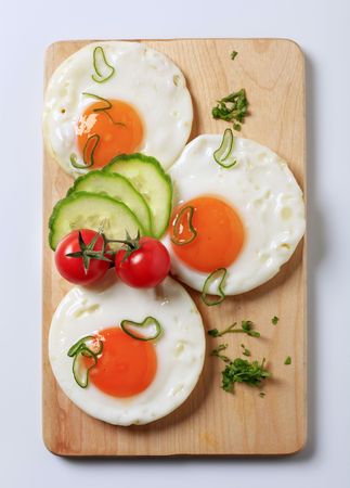 Three fried eggs and fresh vegetables on a cutting boardの写真素材