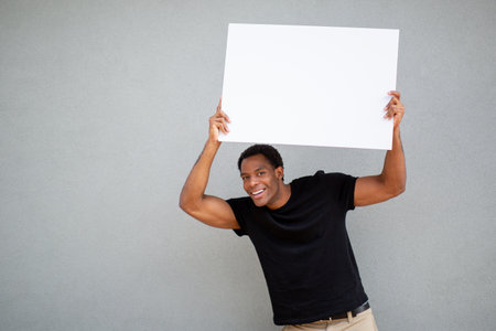 Smiling man wearing a black t-shirt and beige pants holding a blank white sign above his head while standing in front of a grey wallの写真素材