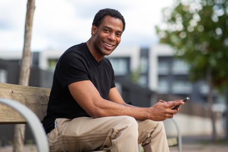 Man sitting on a wooden bench outdoors, smiling and holding a smartphone in his handsの写真素材