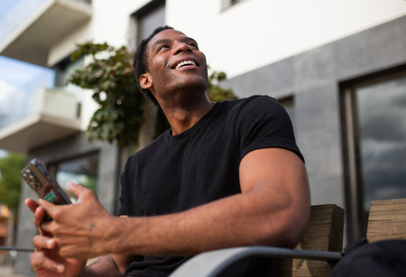 Man wearing a black t-shirt sitting on a bench, smiling and looking up while holding a smartphoneの写真素材