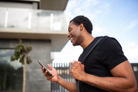 Side view of a man in a black t-shirt smiling and using his smartphone while walking outsideの写真素材