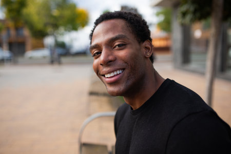 Close-up of a man sitting outdoors, smiling at the camera. Blurred background with urban elements and trees, creating a casual atmosphereの写真素材