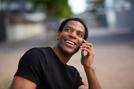 A man in a black t-shirt smiling and talking on a smartphone in an outdoor urban setting, with a blurred backgroundの写真素材