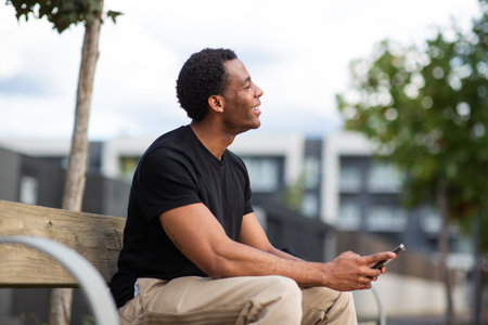 Man sitting on a wooden bench outdoors, looking up, smiling and holding a smartphone in his handsの写真素材