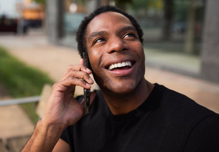 Close-up of a man wearing a black t-shirt sitting on a bench, smiling while talking on a smartphoneの写真素材