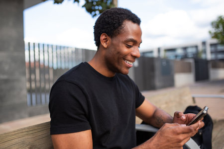 Man sitting on a wooden bench outdoors, smiling as he uses a smartphoneの写真素材