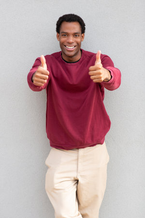 A man giving two thumbs up, wearing a red sweater and beige pants, standing against a gray wallの写真素材