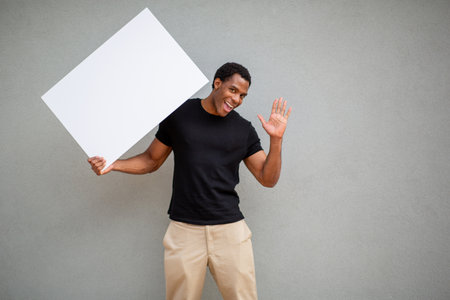A man in a black t-shirt holds a large white poster board in one hand while waving with the other, standing in front of a grey wallの写真素材