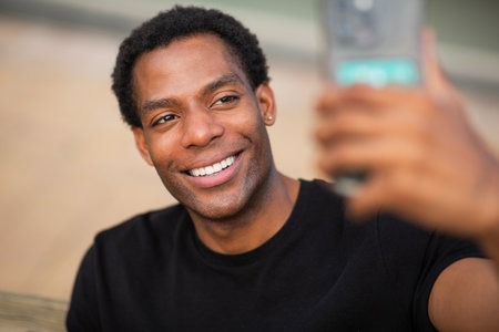 A close-up shot of a man in a black t-shirt smiling while taking a selfie with a smartphoneの写真素材