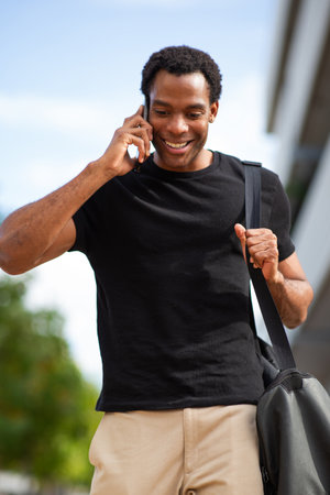 A man wearing a black t-shirt and beige pants talking on his phone while walking outdoors with a shoulder bagの写真素材