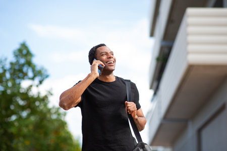 Man in a black t-shirt walking outside, talking on a smartphone and carrying a black shoulder bagの写真素材