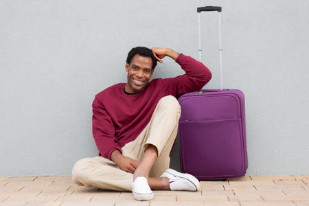 A man in a burgundy sweatshirt and beige pants sitting on the ground next to a purple suitcase, looking at the camera and smilingの写真素材