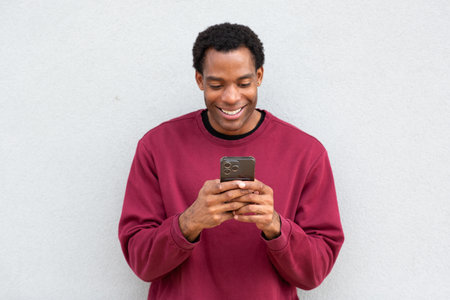 Man in a red sweatshirt standing against a plain wall, looking down at his smartphone and smiling while using both hands to hold itの写真素材