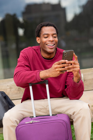 Smiling man sitting on a wooden bench, holding a smartphone with a purple suitcase in front of himの写真素材