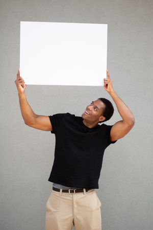 Man wearing a black t-shirt and beige pants holding a blank white sign above his head while looking up at it, standing against a grey wallの写真素材