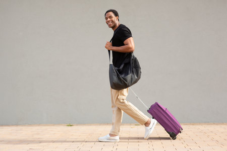 Man in casual clothes walking on sidewalk, pulling a purple suitcase and carrying a black shoulder bagの写真素材