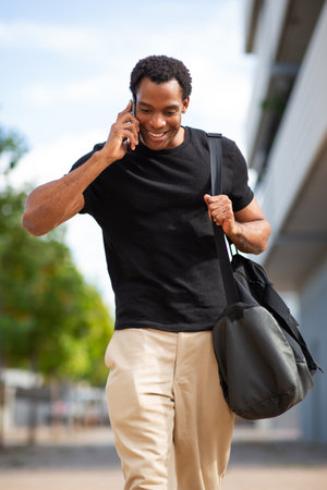 A man dressed in a black shirt and beige pants walking outdoors and talking on his phoneの写真素材