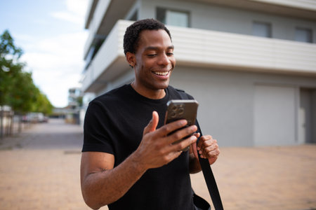 Young man in a black t-shirt smiling and looking at his smartphone while walking outsideの写真素材