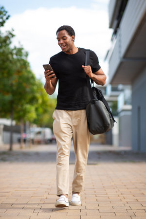 A man wearing a black t-shirt and beige pants walking outdoors while looking at his smartphone, carrying a black shoulder bagの写真素材