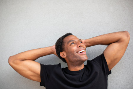 Close-up of a man laughing with his arms behind his head, standing against a grey wallの写真素材