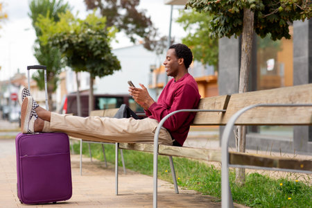 Man sitting on a wooden bench with legs resting on a purple suitcase, using a smartphoneの写真素材