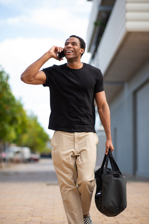 A man in a black t-shirt and beige pants walking outside, holding a black bag in one hand and talking on his smartphone with the otherの写真素材