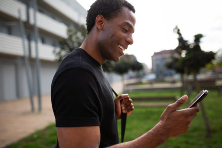 Side view of an African American man in a black t-shirt holding a smartphone and smiling while...