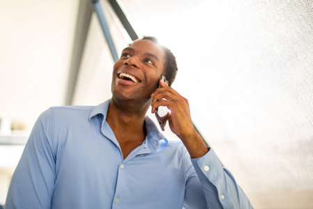 A man in a blue shirt laughing as he talks on the phone in an outdoor settingの写真素材