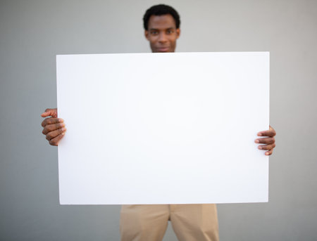Man in a black t-shirt and beige pants holding a blank white sign with both hands while standing against a grey wallの写真素材