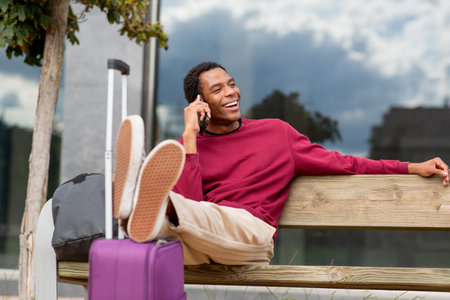 Man lying back on a wooden bench outdoors, talking on his smartphone and smiling. A purple suitcase is next to him, along with a black backpack, with a cloudy sky in the backgroundの写真素材