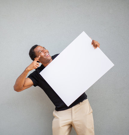A man in a black shirt holds a blank white poster board while pointing up to the side, standing in front of a grey wallの写真素材