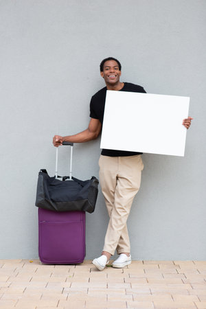 Man in casual clothing standing against a wall, holding a blank white sign, with a purple suitcase and black shoulder bag beside himの写真素材